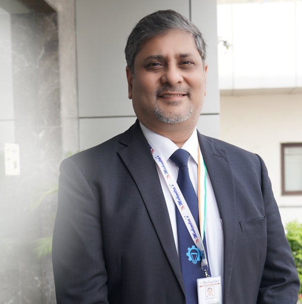 Mr. Naveen Kumar Jain, in a suit and tie, stands before a building.